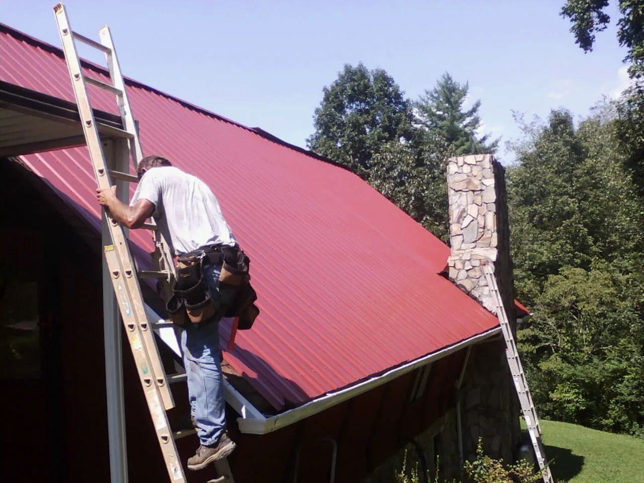 Roofer on a ladder beside a red metal roof and stone chimney — Soffit & Fascia Repair by Roofing Brothers in Cove
